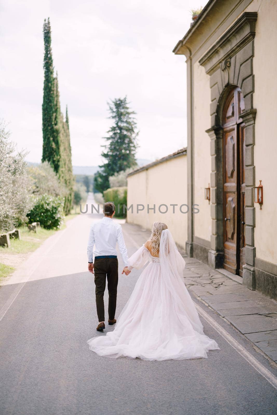 Beautiful bride and groom walking hand in hand away from the camera outside of old villa in Italy, in Tuscany, near Florence by Nadtochiy