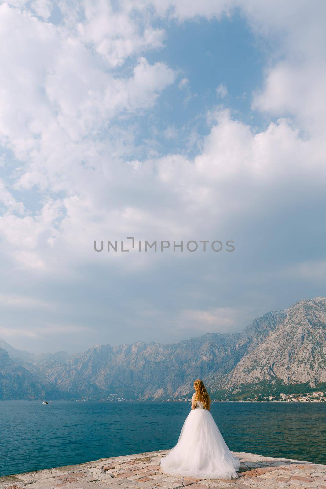 A beautiful blonde bride in a puffy white dress stands on a pier in the Bay of Kotor . High quality photo