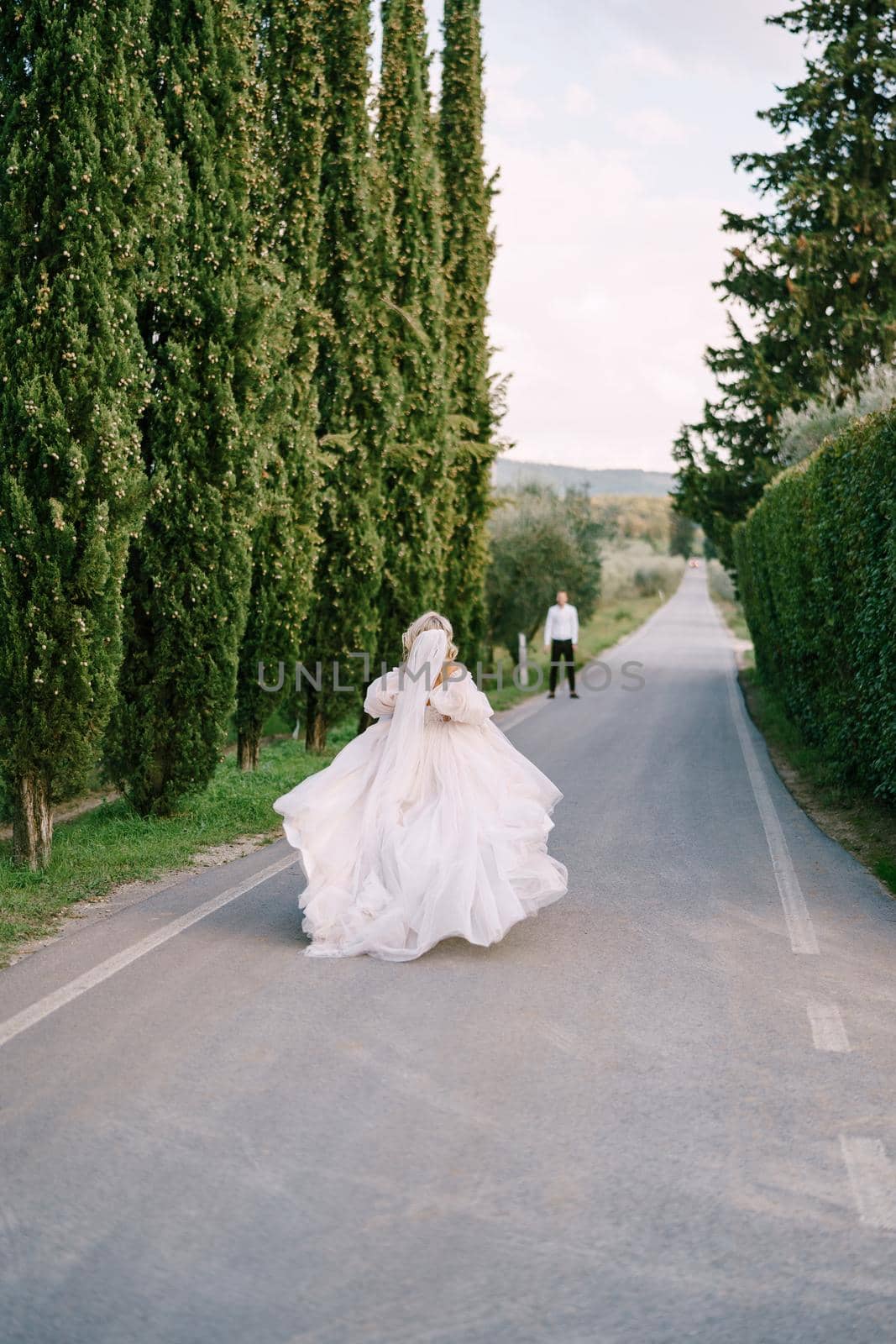 Wedding at an old winery villa in Tuscany, Italy. The bride runs on the way to the groom. A row of cypresses along the road.