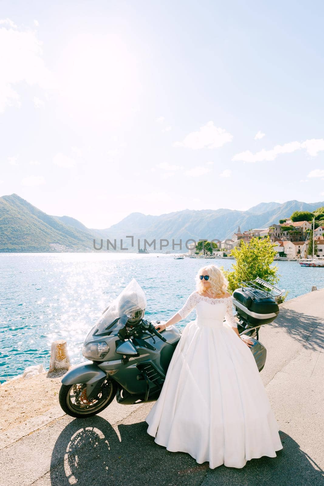 A bride in sunglasses and a wedding dress stands near a motorcycle on the pier in the old town of Perast . High quality photo