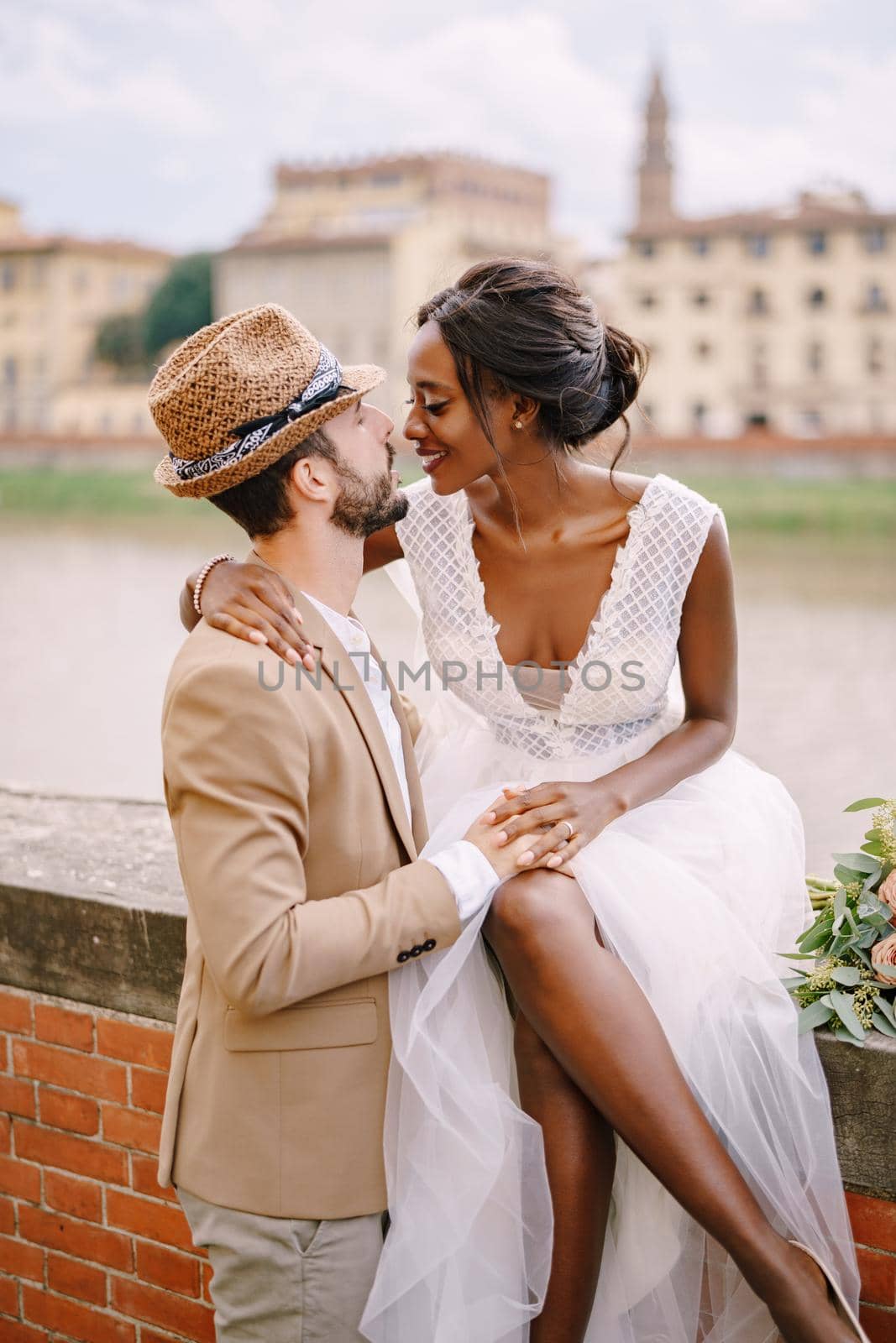 Wedding in Florence, Italy. Interracial wedding couple. An African-American bride is sitting on a brick wall and Caucasian groom is hugging her. Arno River Embankment, overlooking city and bridges by Nadtochiy