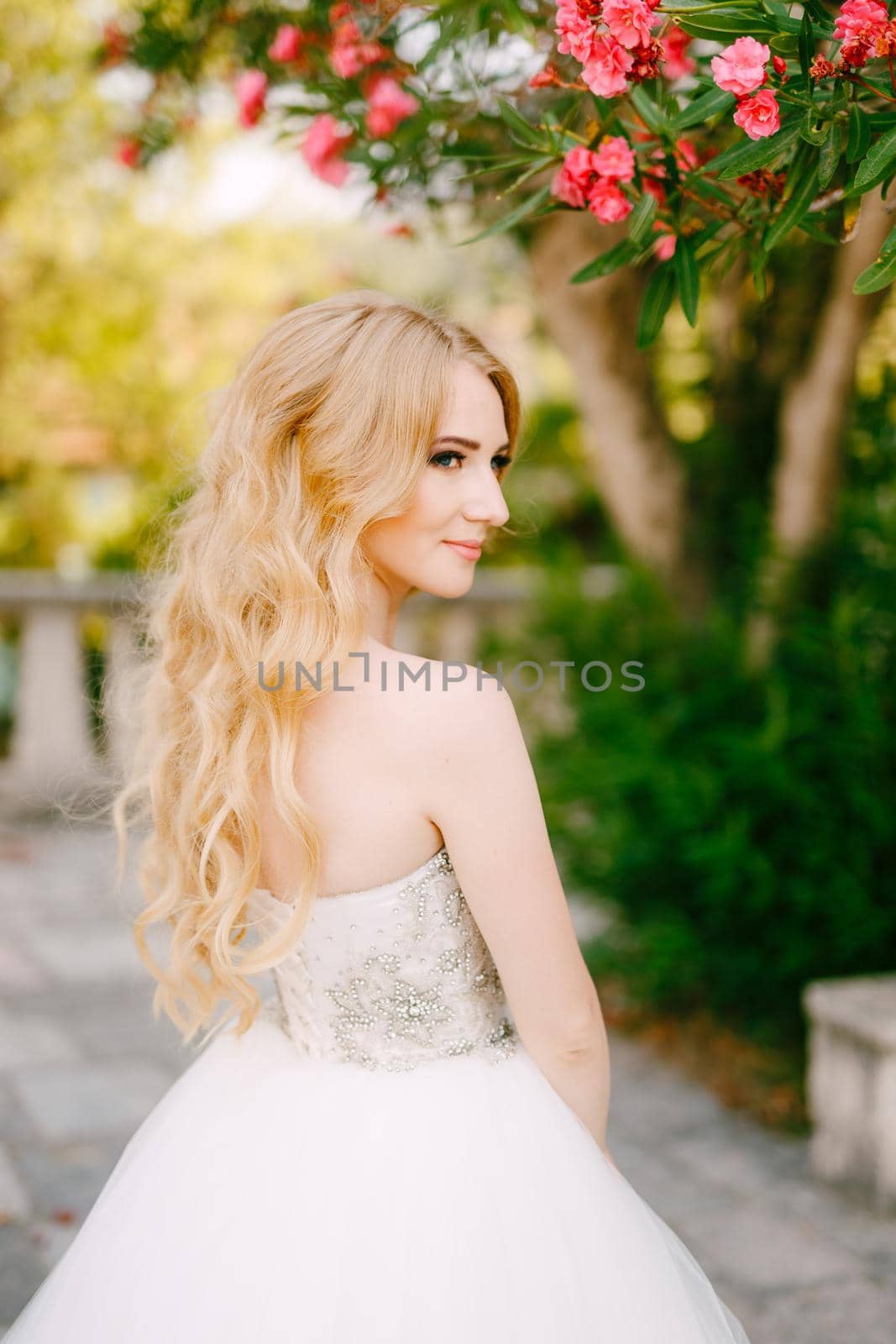 A graceful blonde bride stands under the branches of a blooming oleander near the ancient church in Prcanj by Nadtochiy