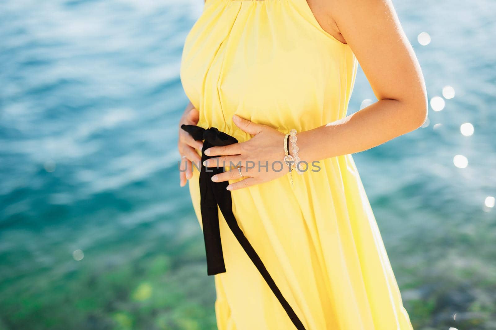 A pregnant woman in a cute bright yellow dress stands by the sea and holds her belly with her hands, close-up. High quality photo