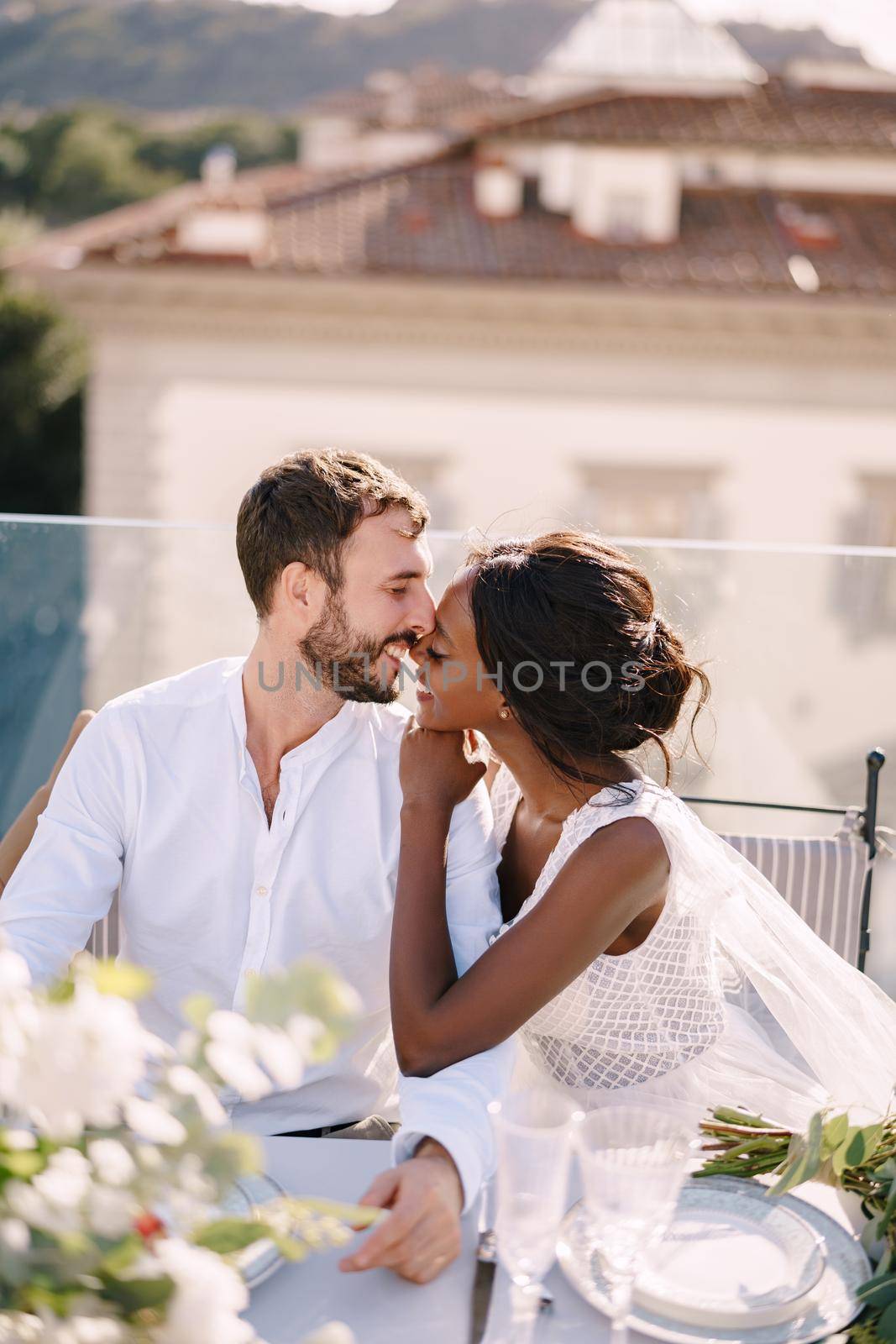 Interracial wedding couple. Destination fine-art wedding in Florence, Italy. African-American bride and Caucasian groom are sitting at rooftop wedding dinner table overlooking the city.