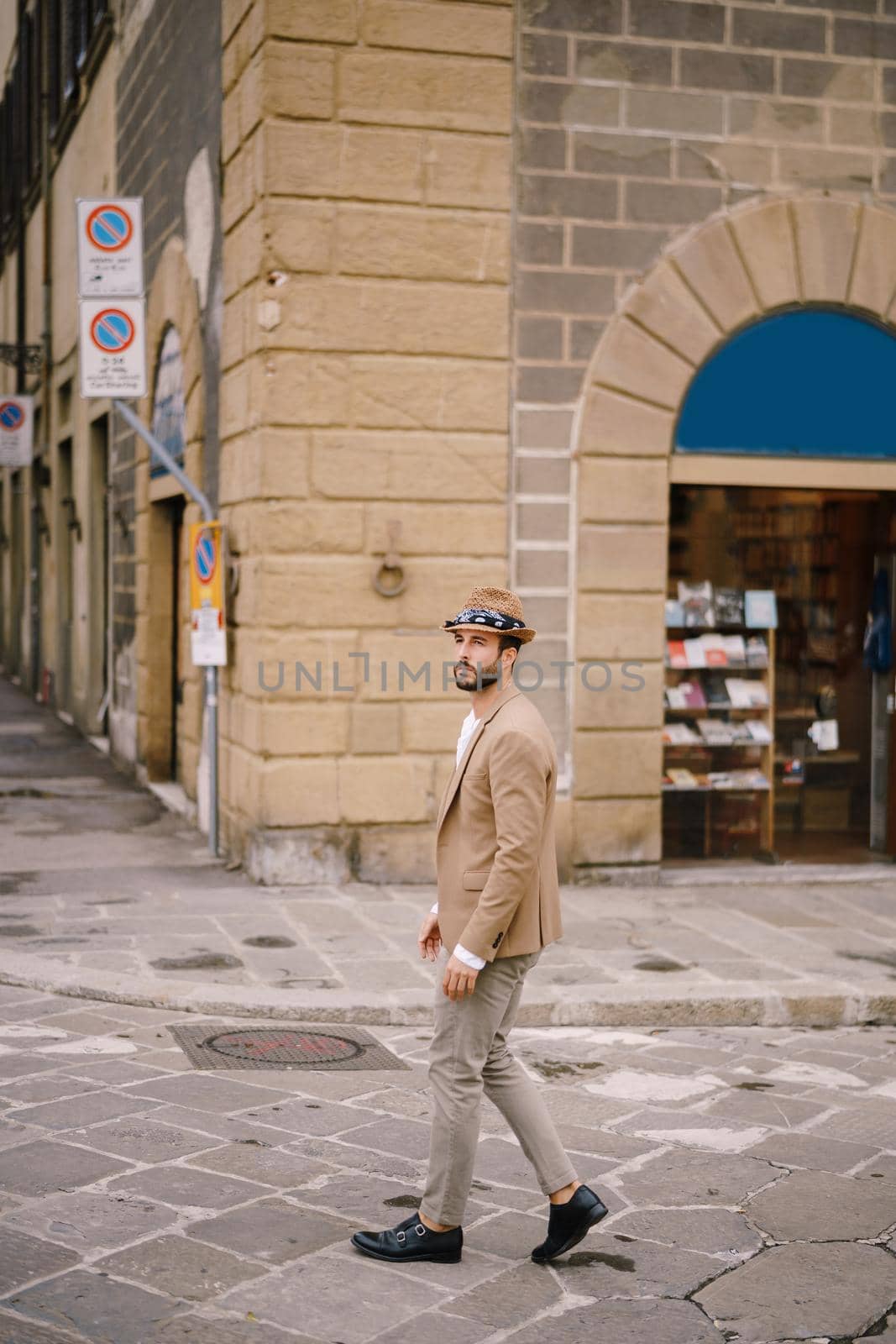 A young guy with a straw hat, a white unbuttoned shirt and a sand jacket, a small beard walks around the city of Florence. by Nadtochiy