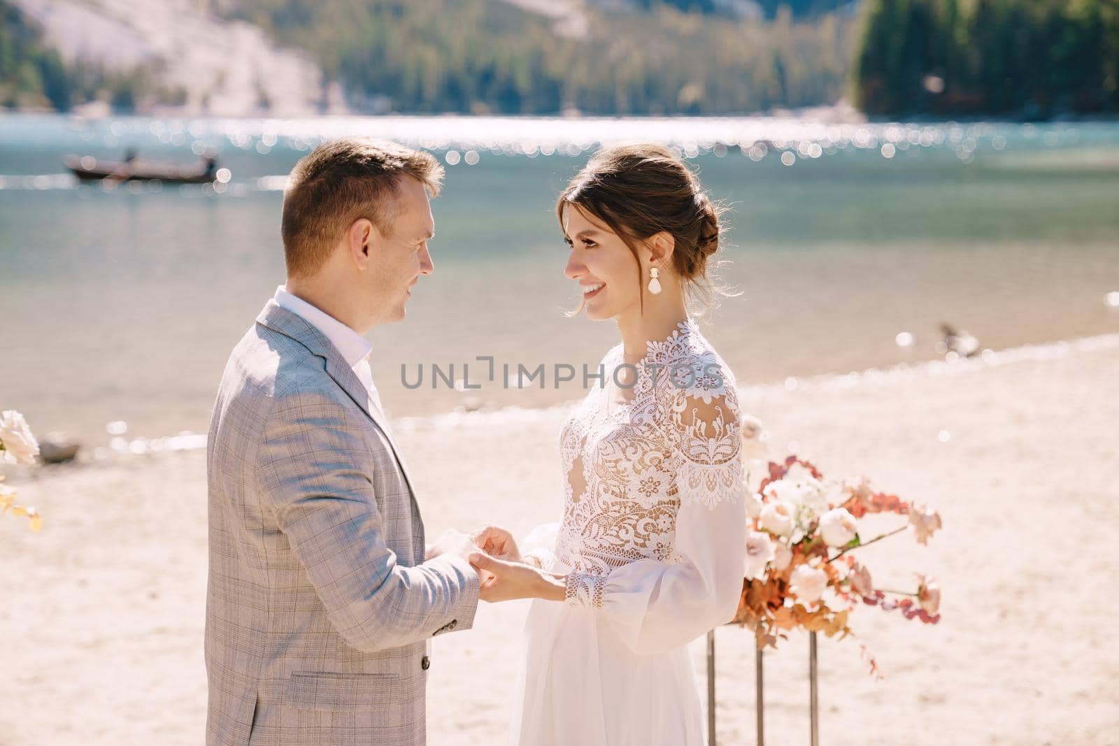 The bride dons a ring to the groom at the venue for the ceremony, with an arch of autumn floral columns, against the backdrop of Lago di Braies in Italy. Destination wedding in Europe, at Braies lake.