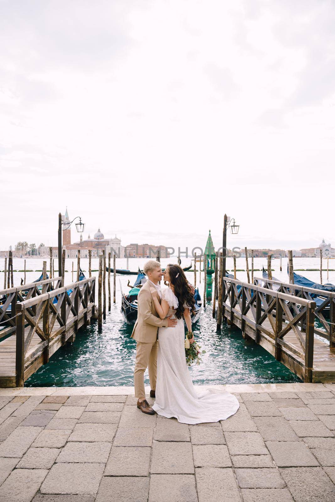 Groom and stand next to the gondola pier, hugging, in Venice, near Piazza San Marco, overlooking San Giorgio Maggiore and the sunset sky. The largest gondola pier in Venice, Italy. by Nadtochiy