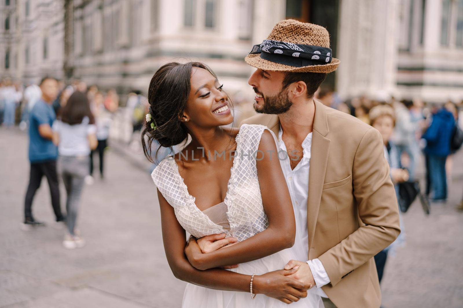 Multiracial wedding couple. Wedding in Florence, Italy. Caucasian groom hugs from behind African-American bride at Piazza del Duomo. by Nadtochiy