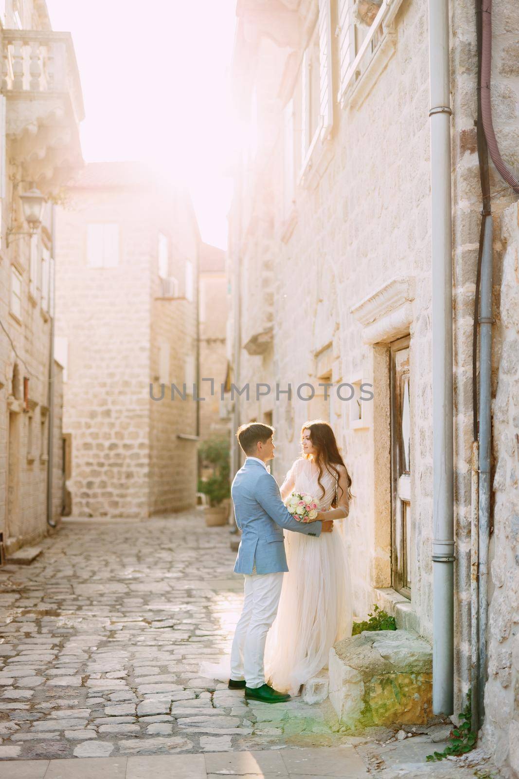 The bride and groom hug on a beautiful old street of Perast near a white stone wall by Nadtochiy