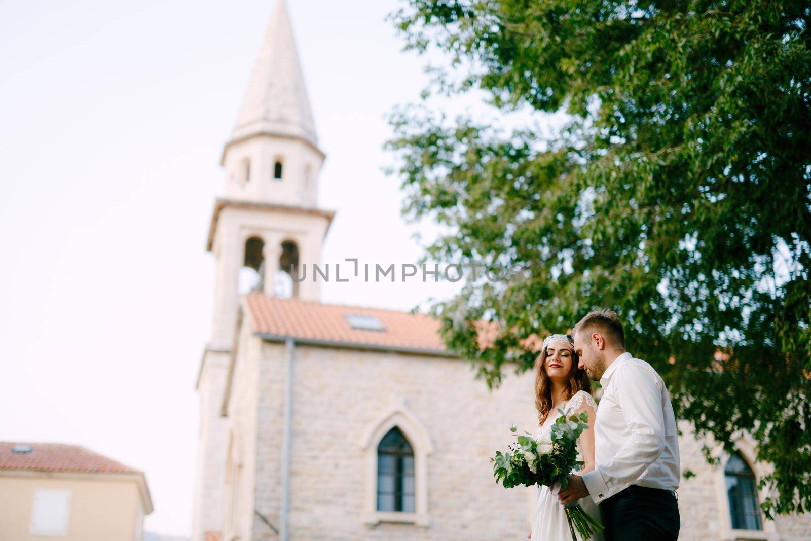 The bride and groom walk through the old town of Budva, the groom gently hugs the bride and holds a bouquet in his hand . High quality photo