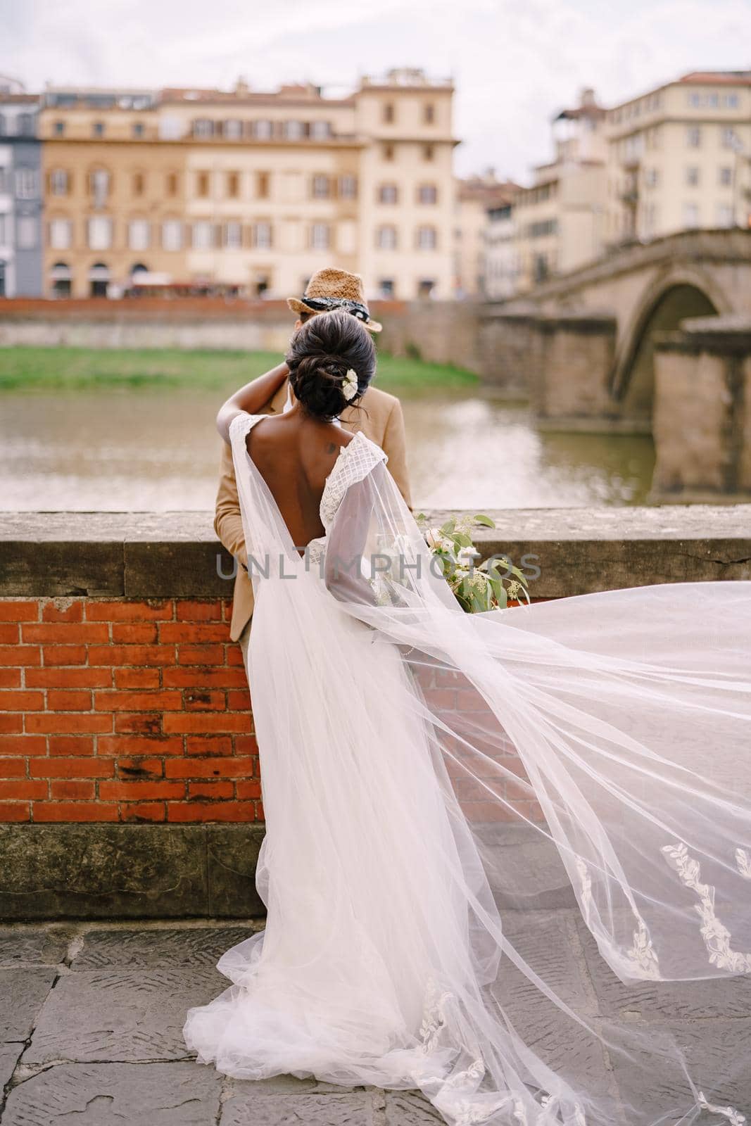 Multiracial wedding couple. Wedding in Florence, Italy. African-American bride and Caucasian groom stand embracing on the embankment of the Arno River, overlooking the city and bridges. by Nadtochiy