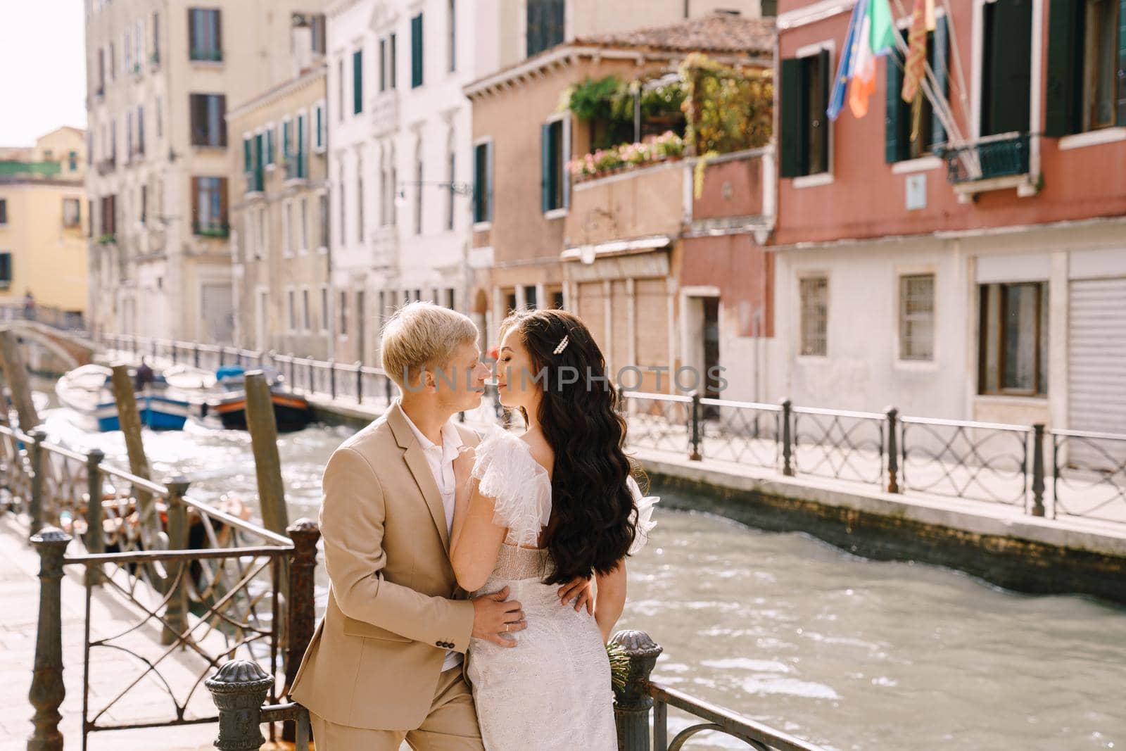 Italy wedding in Venice. Newlyweds stand embracing on the banks of the Venice Canal. The groom hugs the bride by the waist. White wedding dress with small beautiful train and sand-colored men's suit. by Nadtochiy