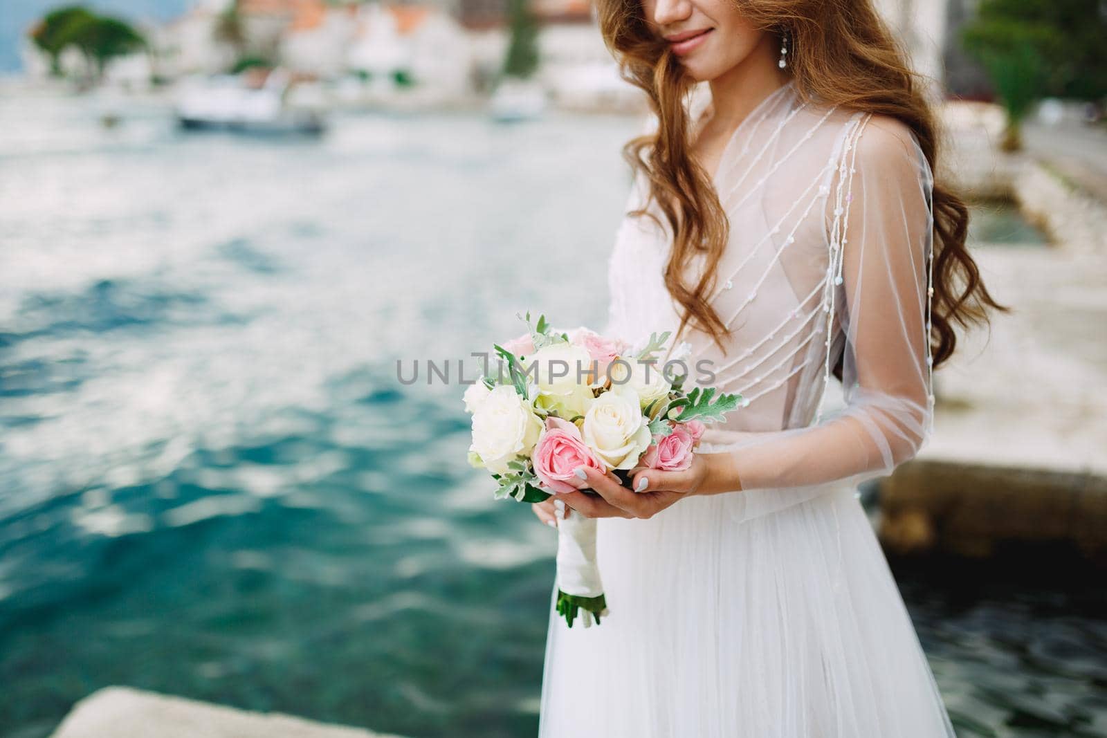 The bride holds a bouquet of roses in her hands and stands on the pier near the old town of Perast . High quality photo