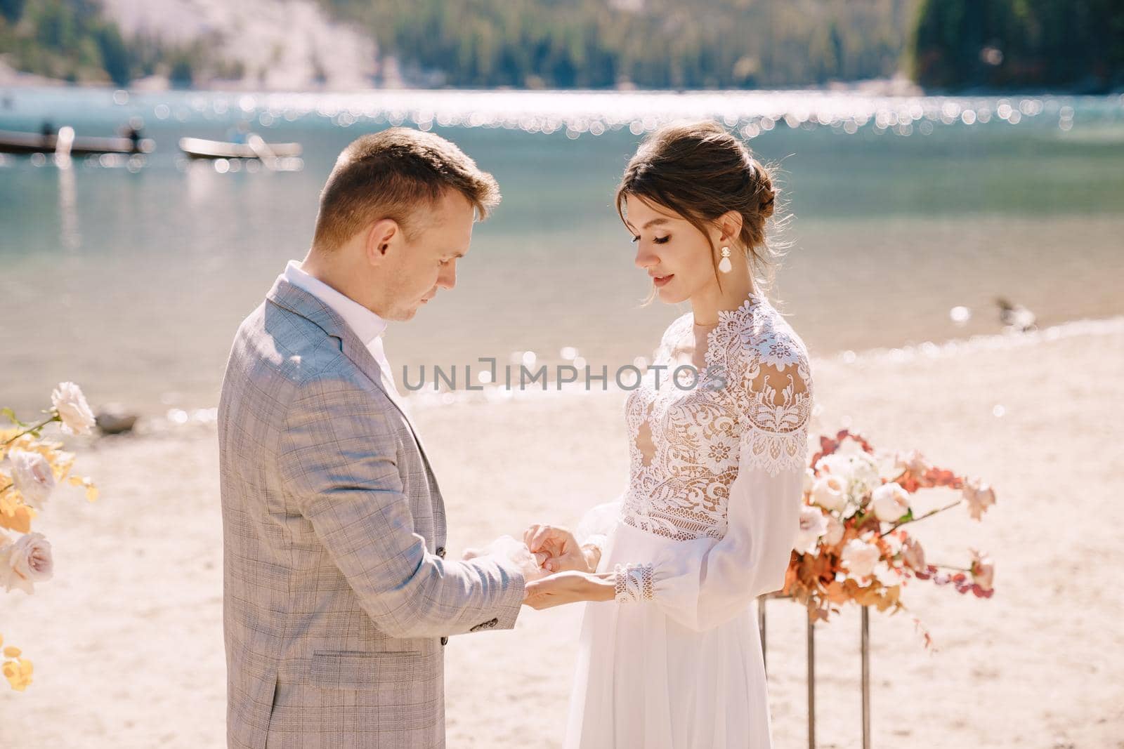 The bride dons a ring to the groom at the venue for the ceremony, with an arch of autumn floral columns, against the backdrop of Lago di Braies in Italy. Destination wedding in Europe, at Braies lake.