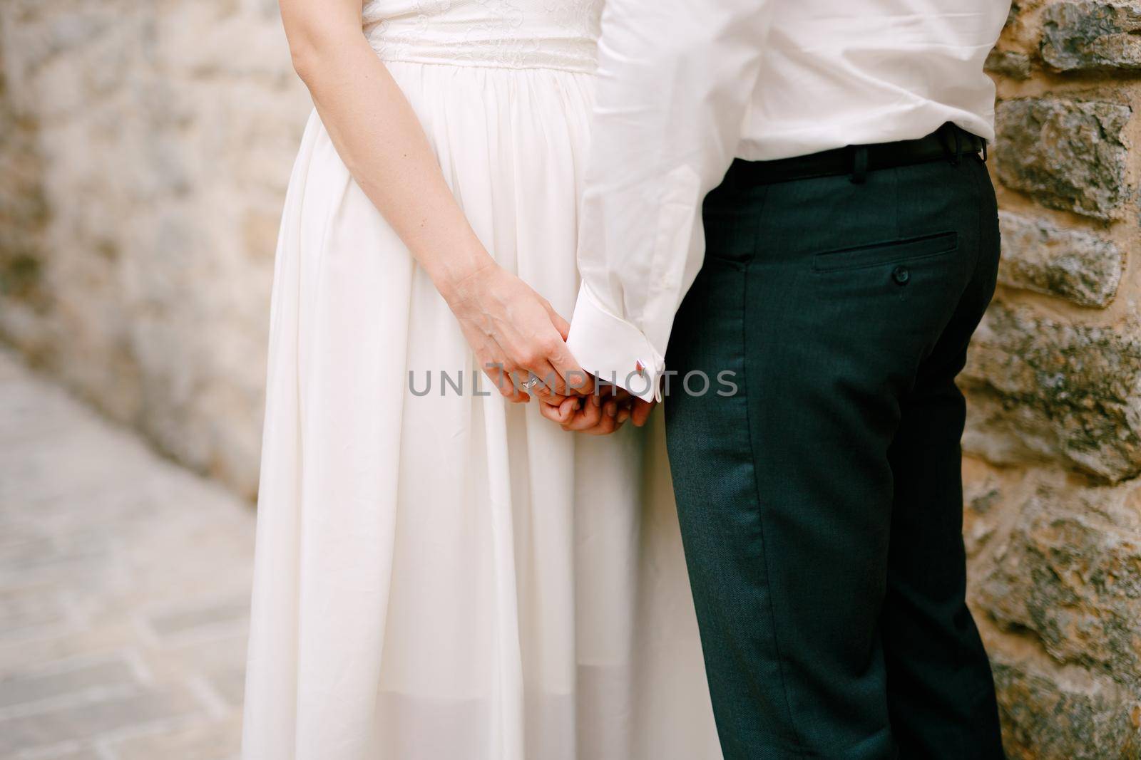 The bride and groom stand near a stone wall and touchingly hold each other's hands, close-up by Nadtochiy