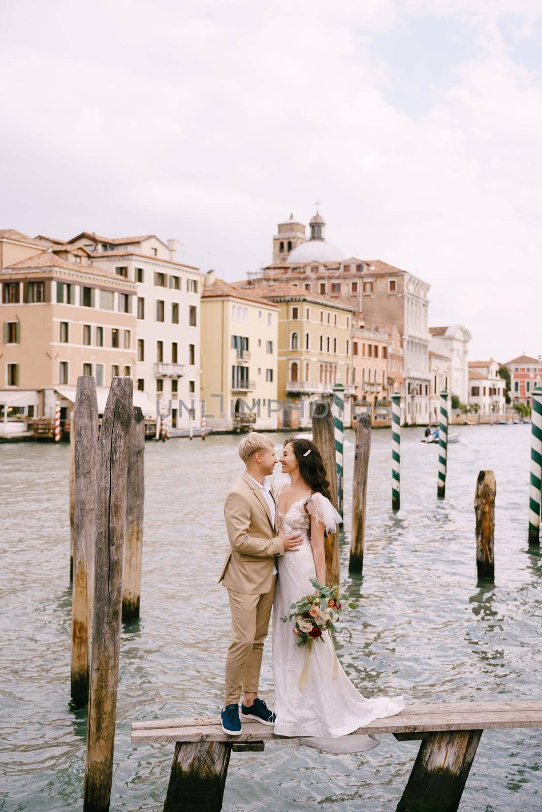 Italy wedding in Venice. The bride and groom are standing on a wooden pier for boats and gondolas, near the Striped green and white mooring poles, against backdrop of facades of Grand Canal buildings. by Nadtochiy