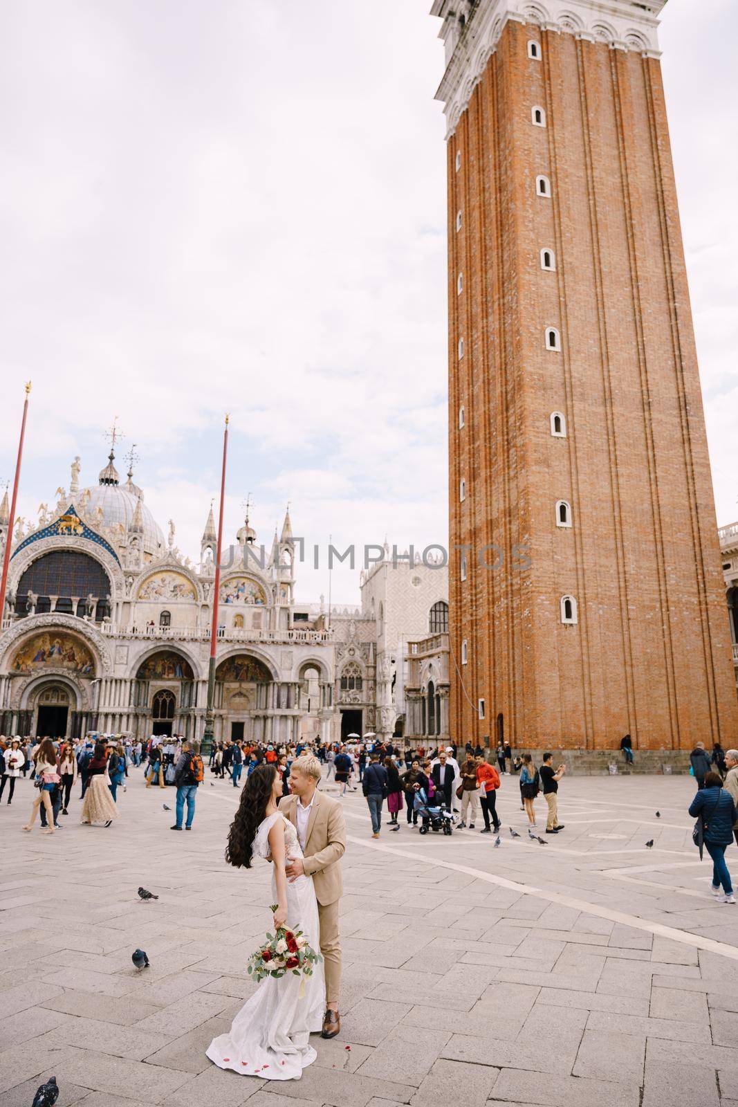 Wedding in Venice, Italy. The bride and groom cuddle in Piazza San Marco, overlooking Campanoil and St. Mark Cathedral, among the crowds of tourists.