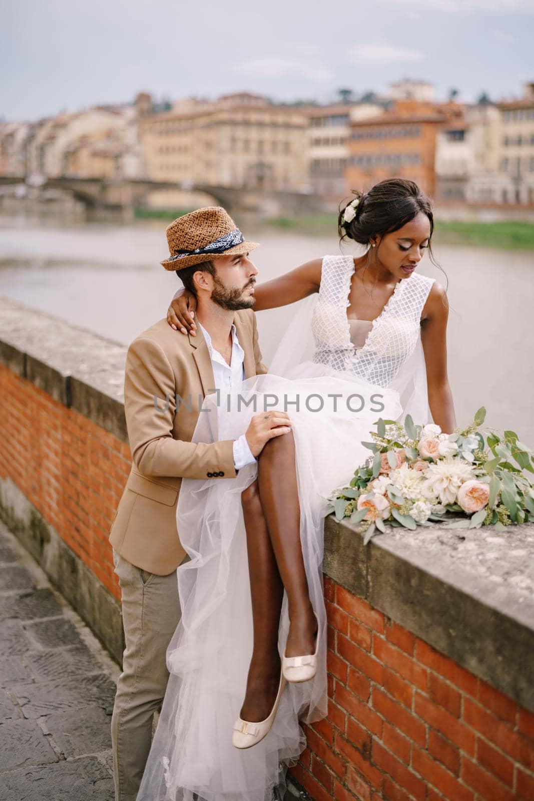 Wedding in Florence, Italy. Multiracial wedding couple. An African-American bride is sitting on a brick wall and Caucasian groom is hugging her. Arno River Embankment, overlooking city and bridges by Nadtochiy