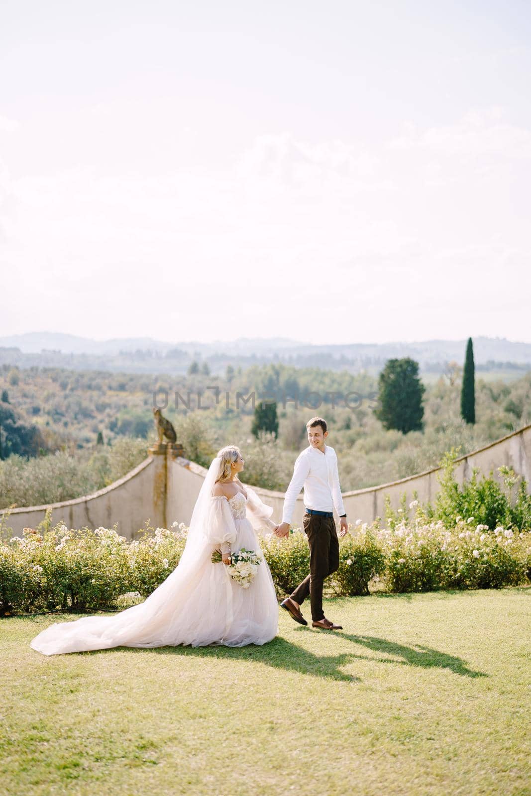 The bride and groom walk in the park. Wedding at an old winery villa in Tuscany, Italy. Round wedding arch decorated with white flowers and greenery in front of an ancient Italian architecture. by Nadtochiy