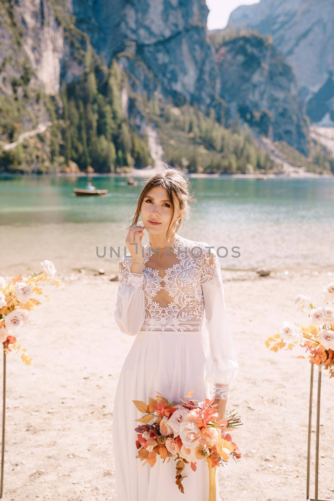 Beautiful bride in a white dress with sleeves and lace, with a yellow autumn bouquet against backdrop of arch for ceremony, at Lago di Braies in Italy. Destination wedding in Europe, at Braies lake.