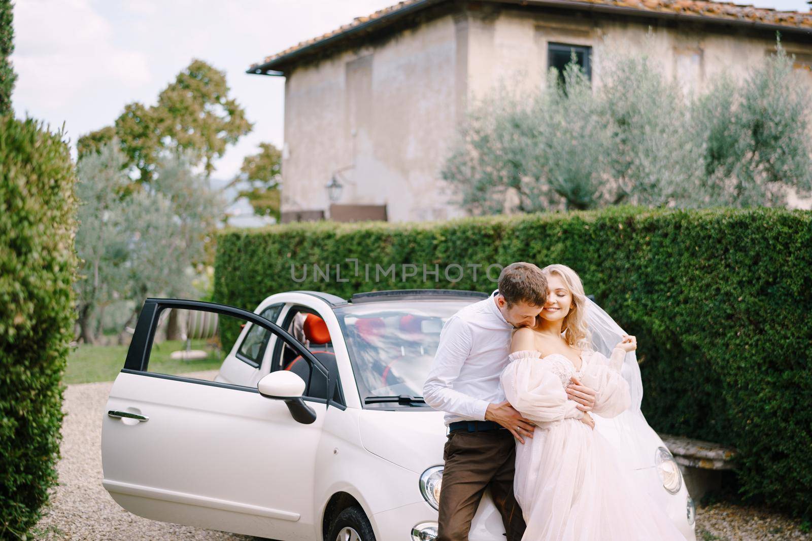 Groom kissing his bride on the dhoulder in front of a convertible at the old villa in Italy, in Tuscany, near Florence