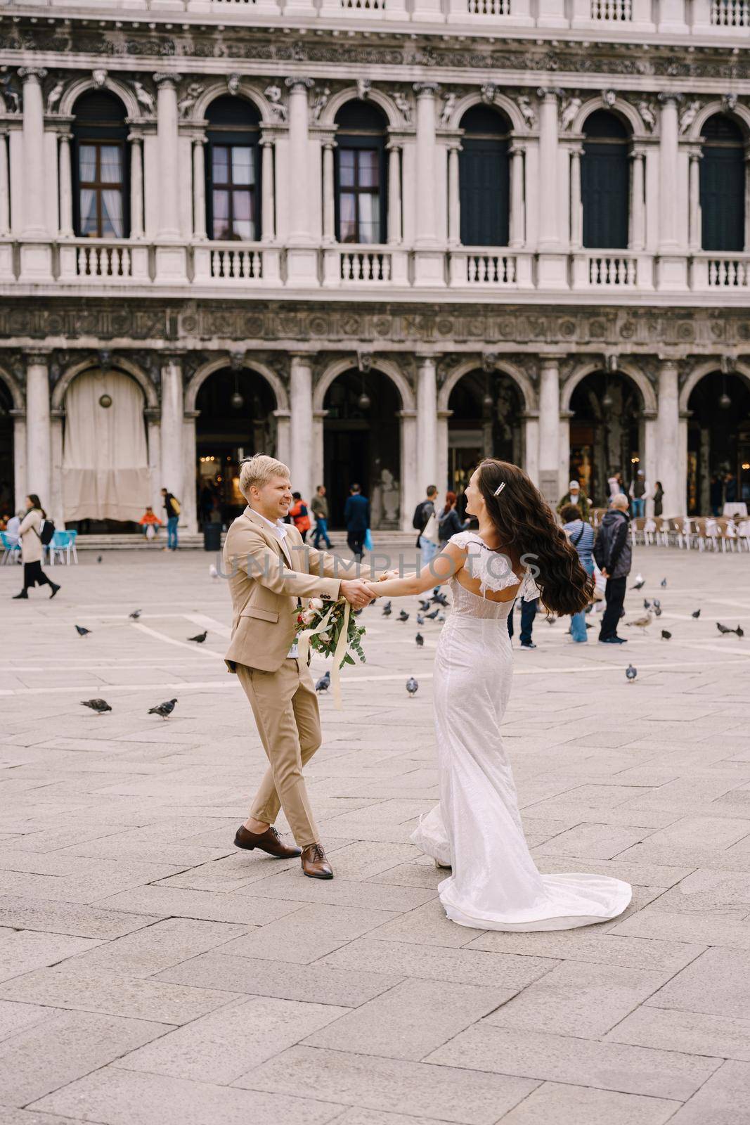Wedding in Venice, Italy. The bride and groom are dancing among many pigeons in Piazza San Marco, against the backdrop of the National Archaeological Museum Venice, surrounded by a crowd of tourists.
