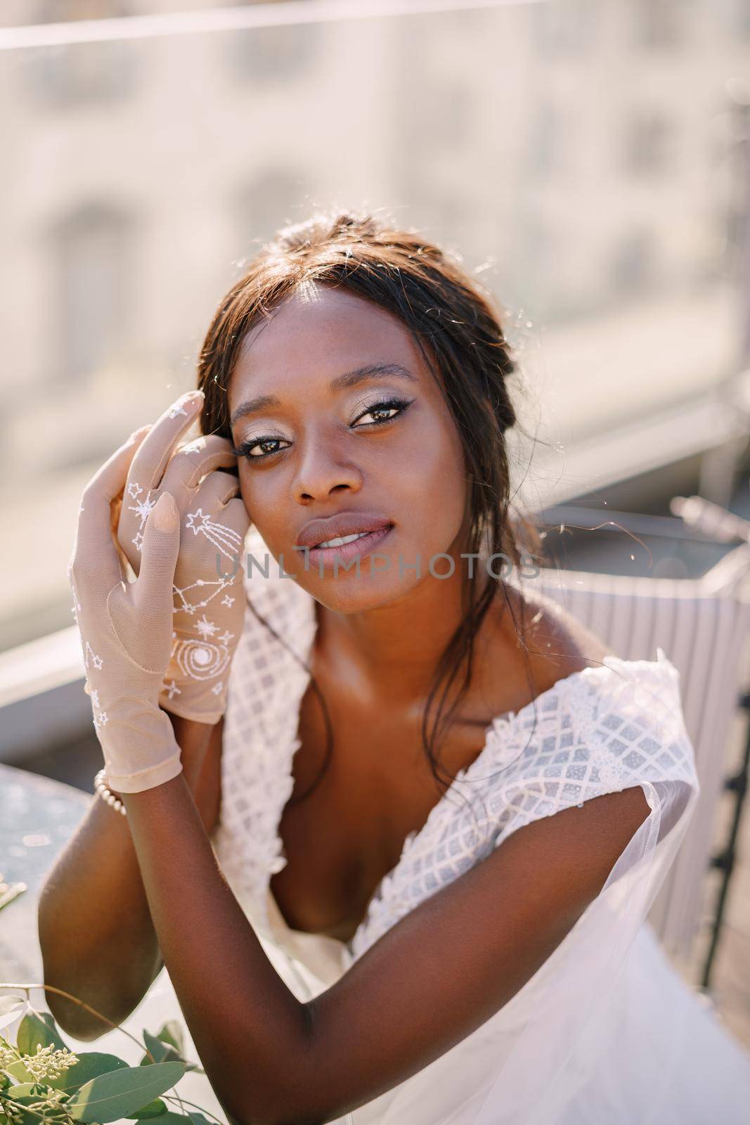 Destination fine-art wedding in Florence, Italy. African-American bride sits at the table, touches his face with her hands in gloves, a bouquet lies on the table.