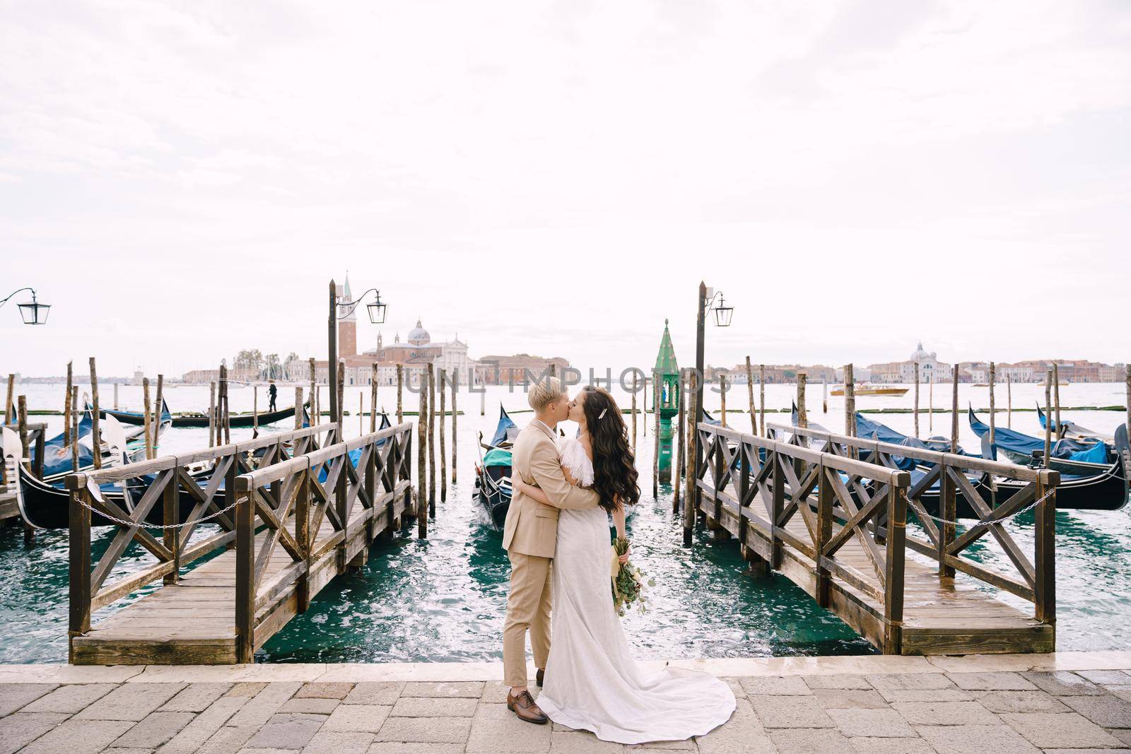 The bride and groom kiss on the gondola pier, hugging, in Venice, near St. Mark's Square, overlooking San Giorgio Maggiore and the sunset sky. The largest gondola pier in Venice, Italy. by Nadtochiy