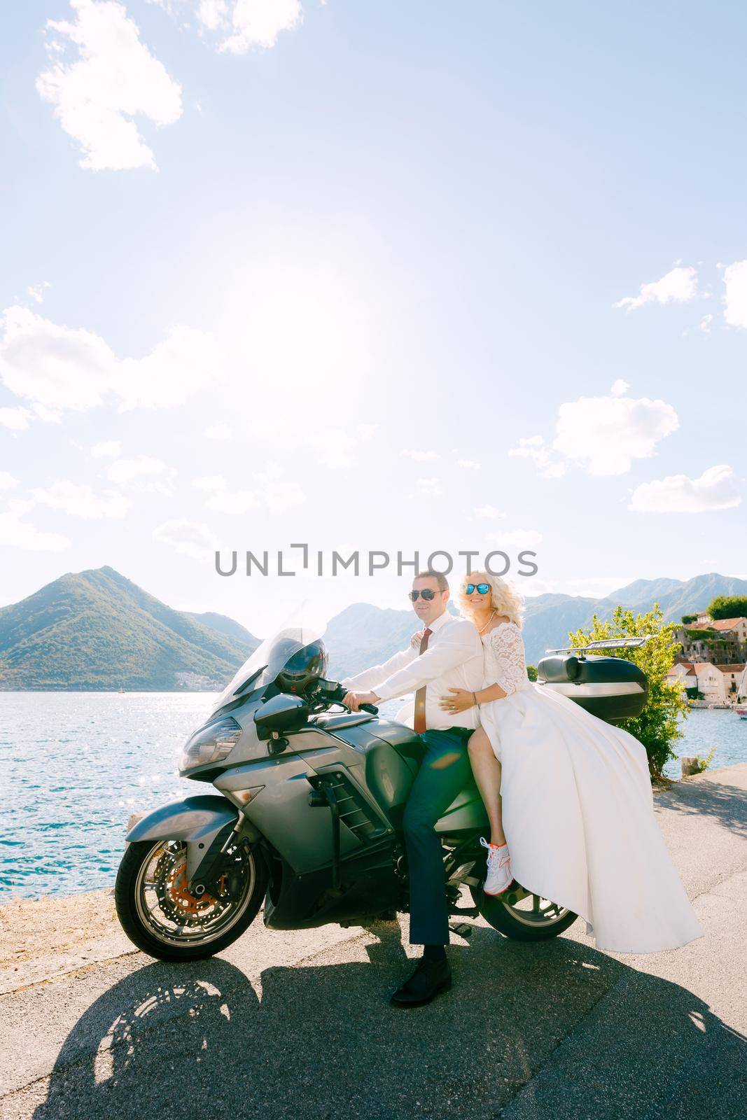The bride and groom sit on a motorcycle on the pier in Perast, the bride hugs the groom . High quality photo