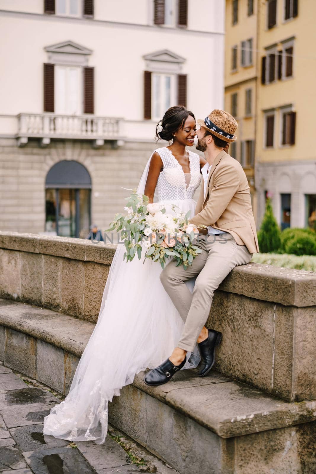 African-American bride in a white dress with a long veil and bouquet, and Caucasian groom in a sand jacket and straw hat. Interracial wedding couple. Wedding in Florence, Italy.