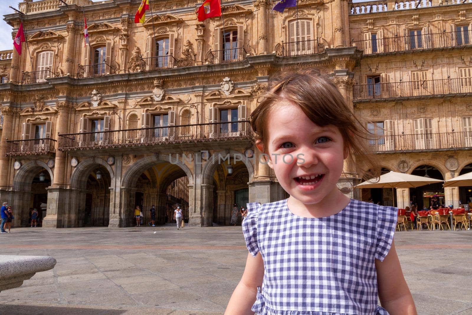 A cute, blue-eyed, brown-haired baby girl smilling at Plaza Mayor de Salamanca on a sunny day