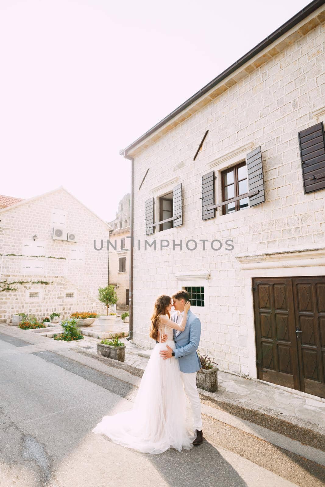 The bride and groom are embracing near the beautiful white house in the old town of Perast by Nadtochiy