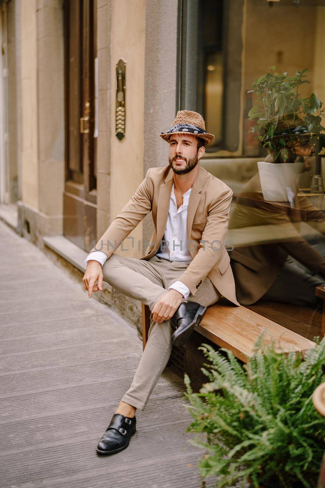 A guy in a sand jacket and gray pants with black shoes, straw hat and a beard is sitting on a street bench.