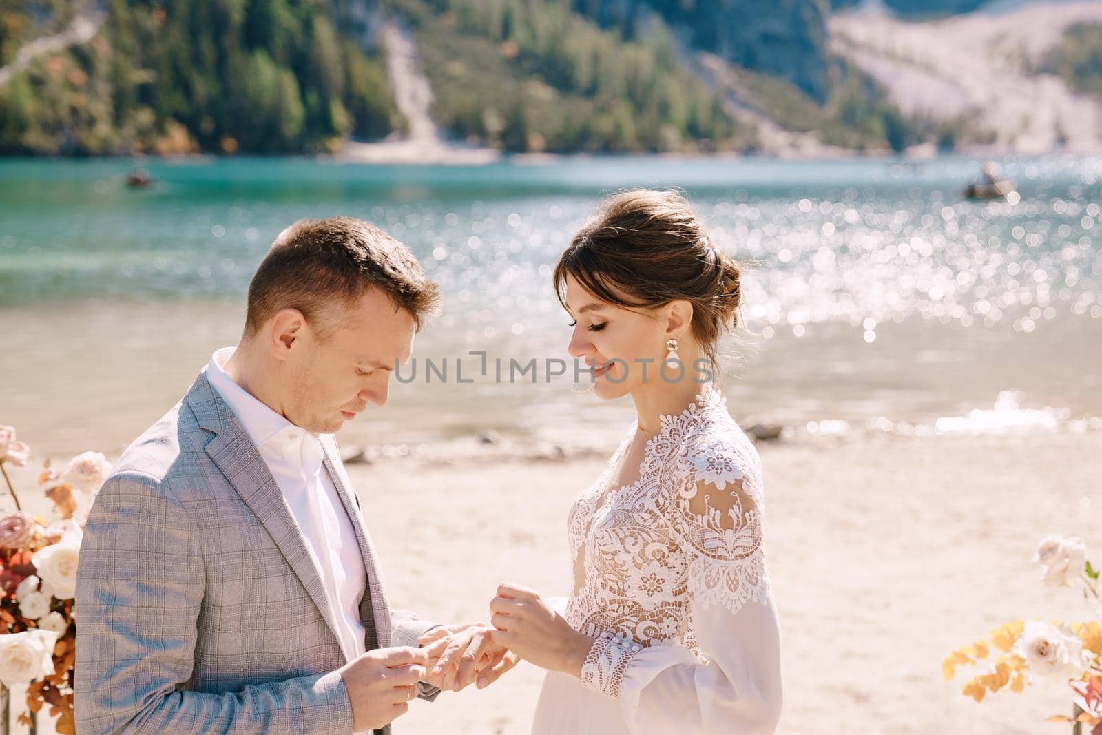 The groom dons a bride's ring, in place for the ceremony, with an arch of autumn floral columns, against the backdrop of Lago di Braies in Italy. Destination wedding in Europe, at Braies lake.
