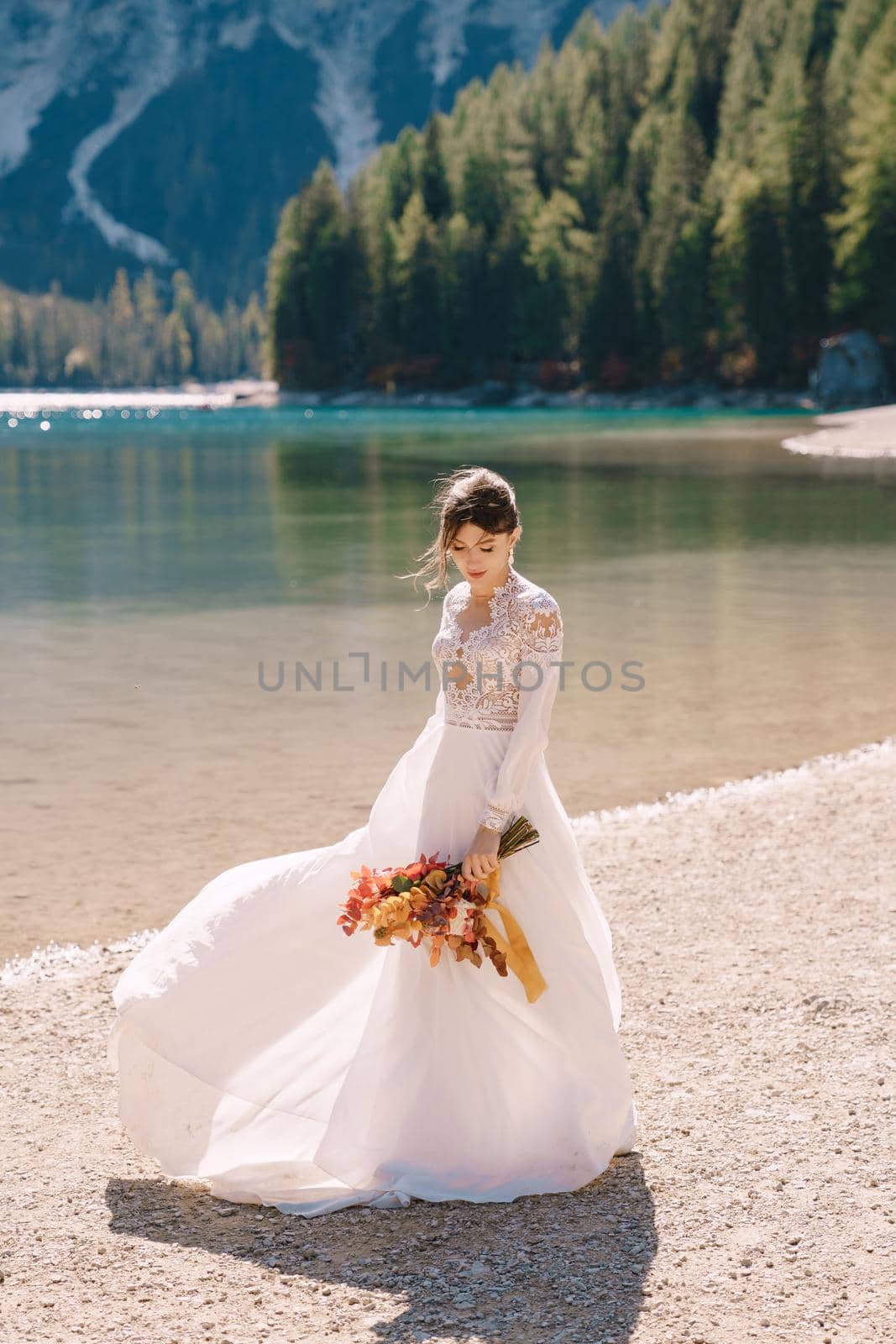 Beautiful bride in a white dress with sleeves and lace, with a yellow autumn bouquet of dried flowers and peony roses, at Lago di Braies in Italy. Destination wedding in Europe, at Braies lake.