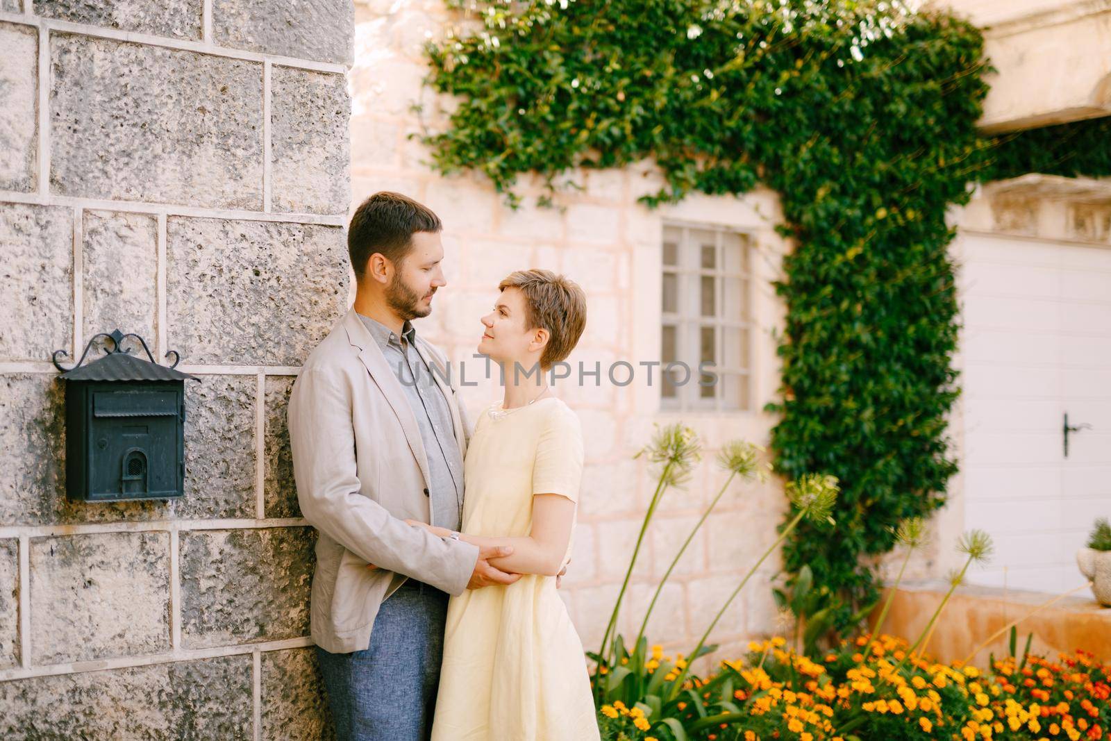 A man and a woman are embracing at the wall of a beautiful house with a window, a liana, flowers and a mailbox . High quality photo