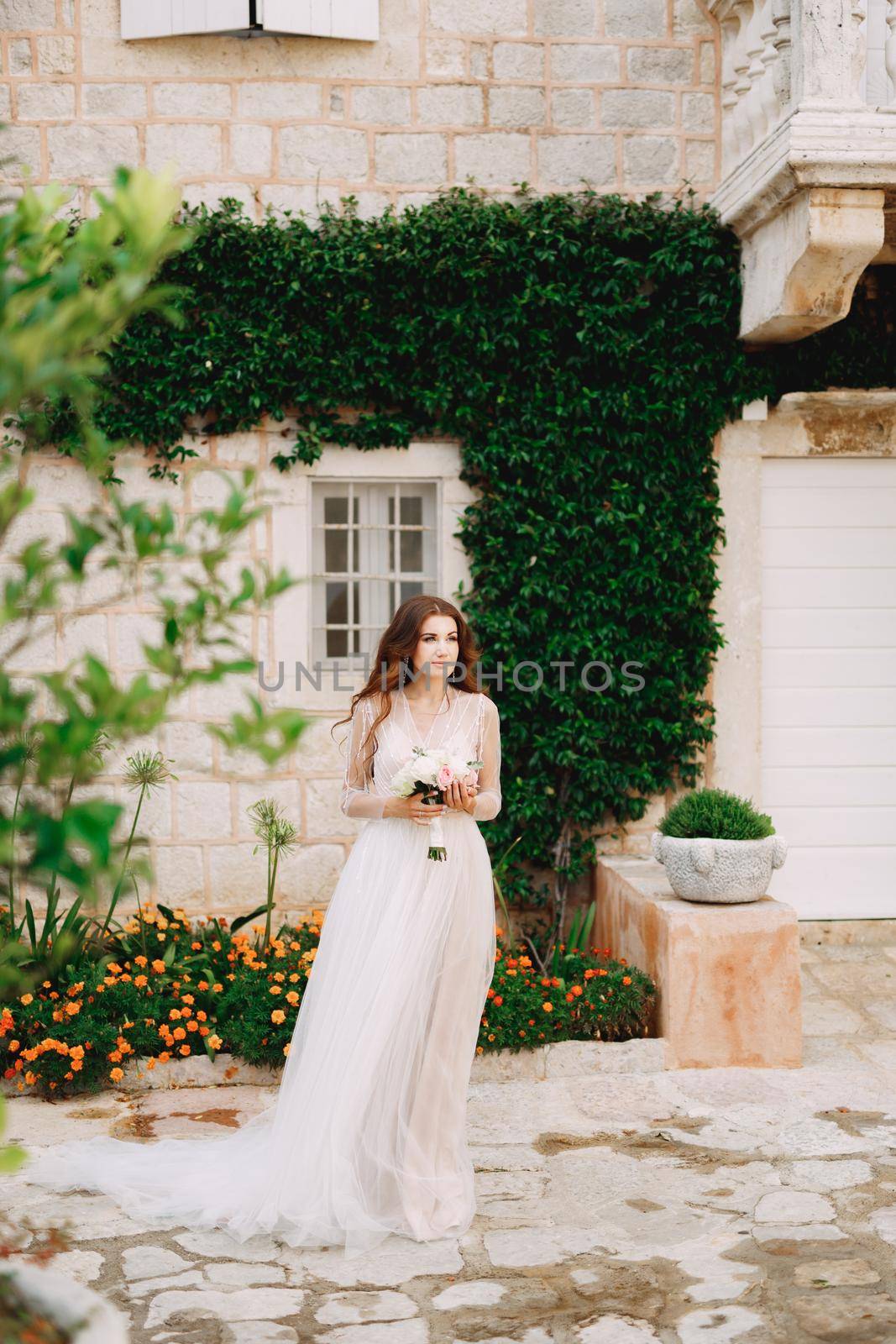 A bride with a bouquet in her hands stands at the wall of a house with a green liana and orange flowers in Perast . High quality photo