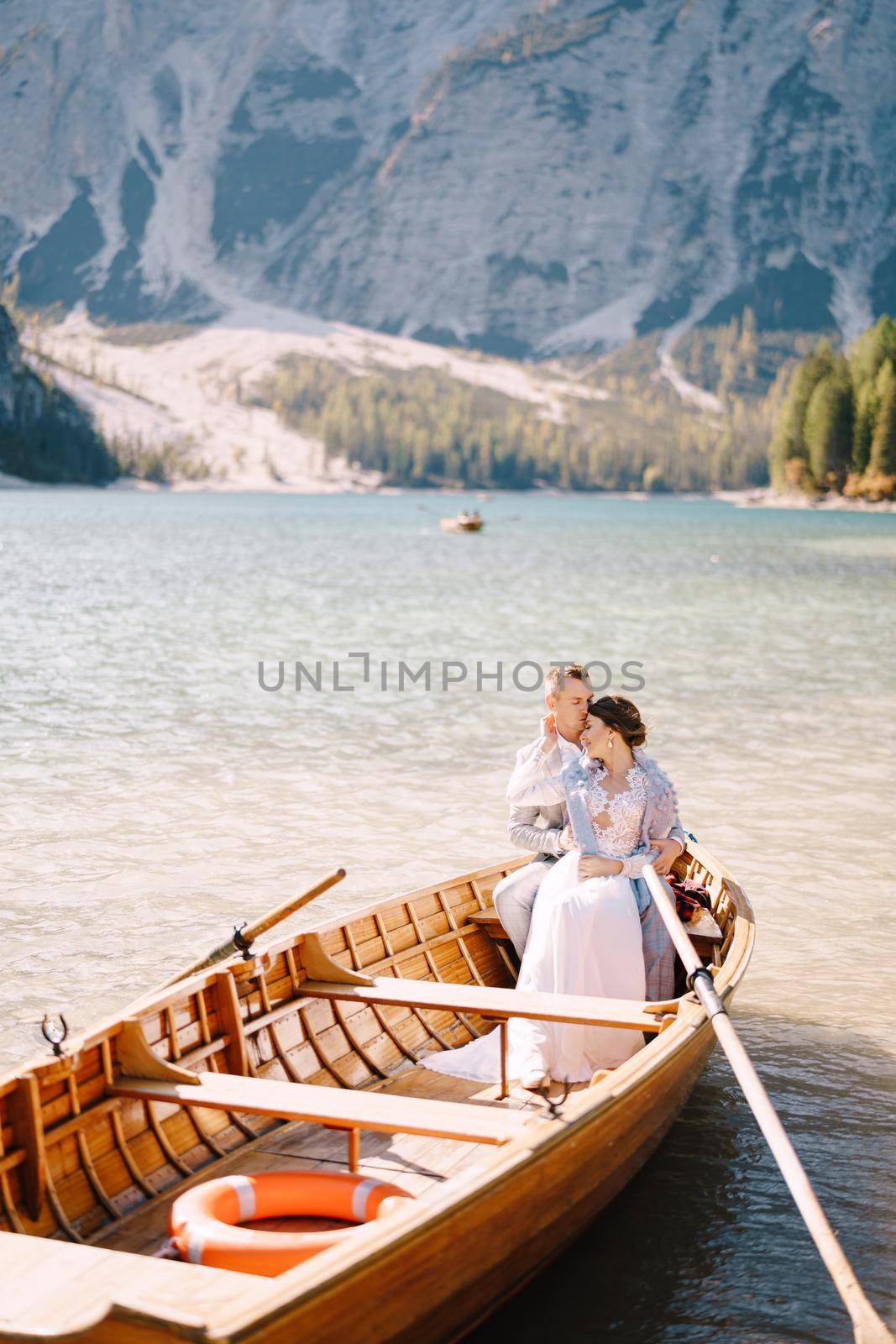 The wedding couple sits in a wooden boat at Lago di Braies in Italy. Newlyweds in Europe, at Braies Lake, in the Dolomites. The groom hugs the bride.