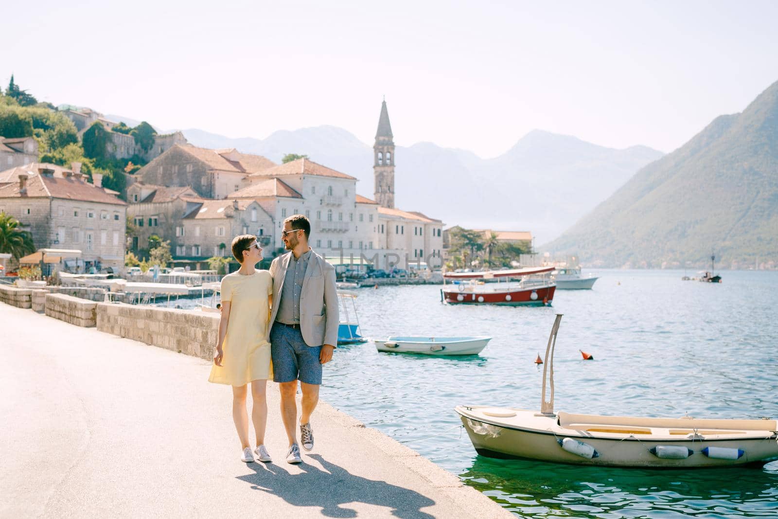 A man and a woman in sunglasses walk hugging each other on the pier near the boats in old town of Perast. High quality photo