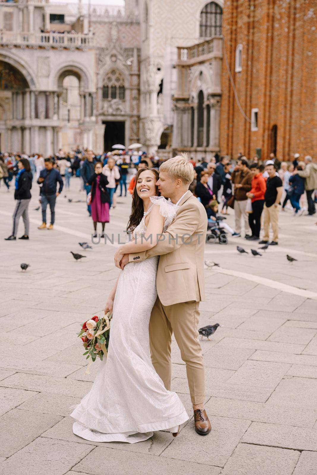 Venice, Italy - 04 october 2019: Wedding in Venice, Italy. The bride and groom are hugging in Piazza San Marco, overlooking Campanila and St. Mark Cathedral, among a crowd of tourists. by Nadtochiy