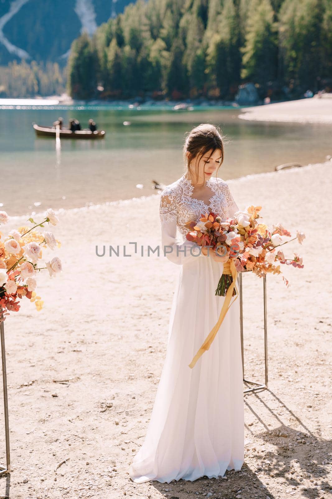 Beautiful bride in a white dress with sleeves and lace, with a yellow autumn bouquet against backdrop of arch for ceremony, at Lago di Braies in Italy. Destination wedding in Europe, at Braies lake.