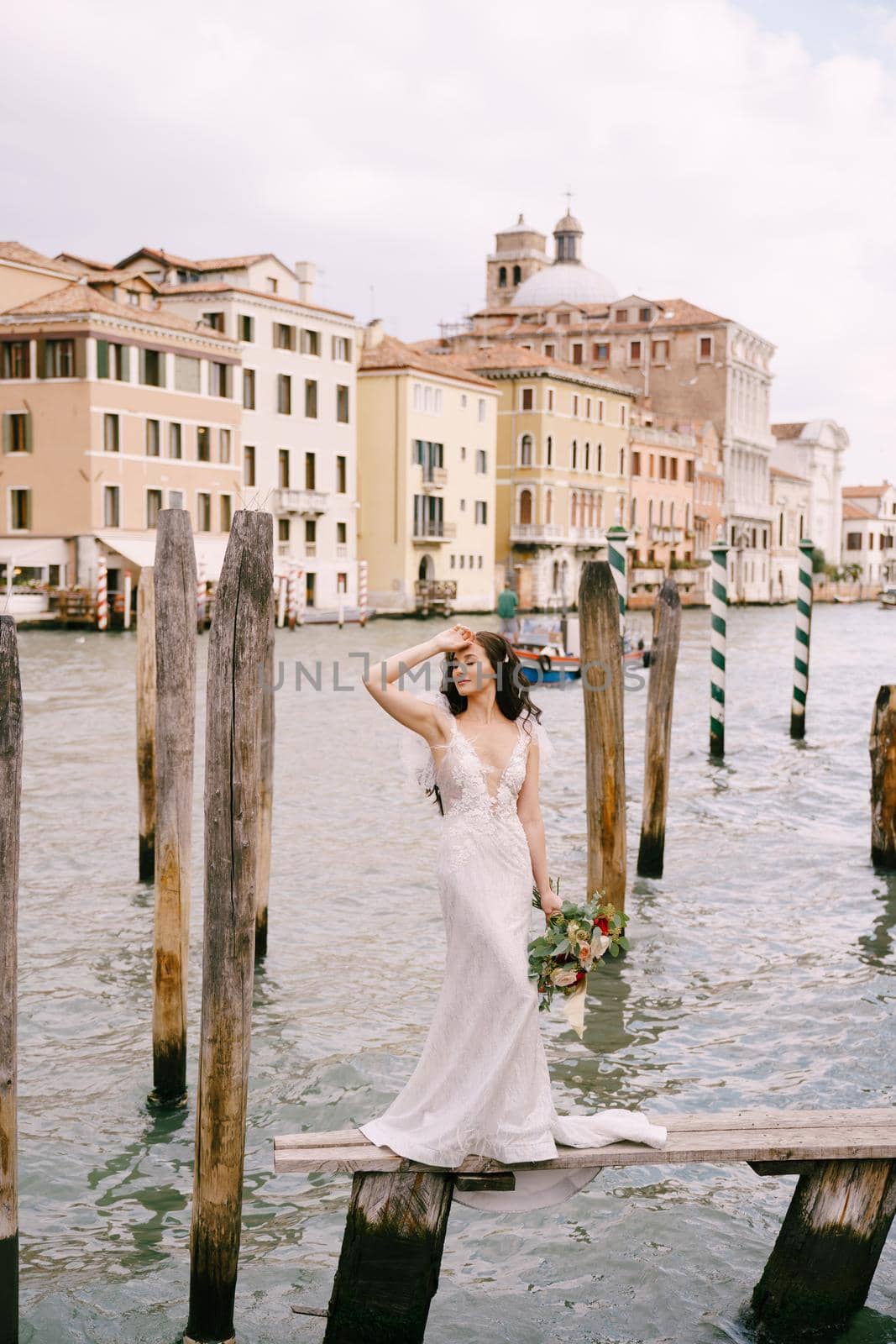 The bride stand on a wooden dock for boats and gondolas, near striped green and white mooring poles, against the facades of the Grand Canal buildings.