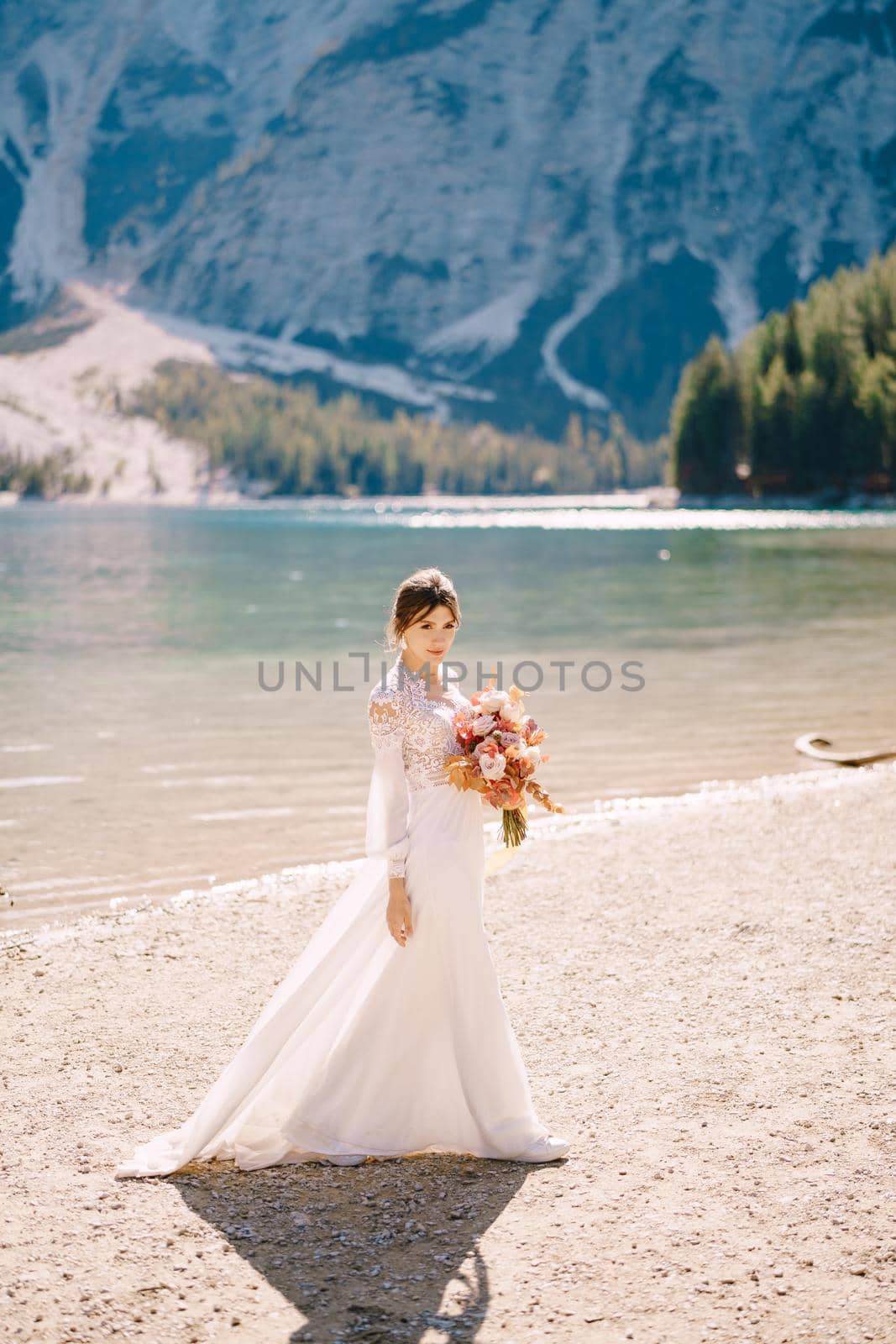 Beautiful bride in a white dress with sleeves and lace, with a yellow autumn bouquet of dried flowers and peony roses, at Lago di Braies in Italy. Destination wedding in Europe, at Braies lake.