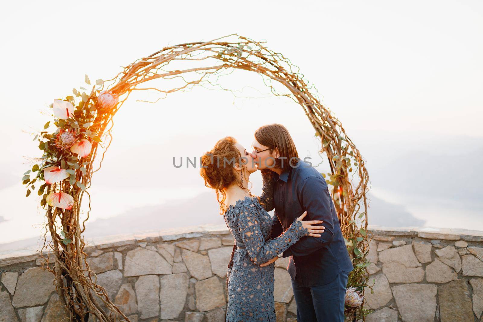 The bride and groom hugging and kissing near the wedding arch on the observation deck overlooking the Bay of Kotor by Nadtochiy