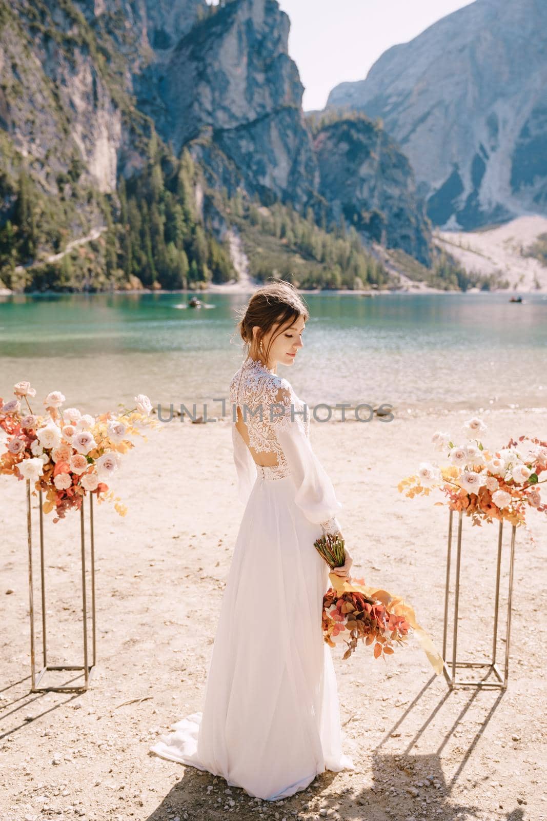 Beautiful bride in a white dress with sleeves and lace, with a yellow autumn bouquet on background of the arch for ceremony, at Lago di Braies in Italy. Destination wedding in Europe, on Braies lake. by Nadtochiy