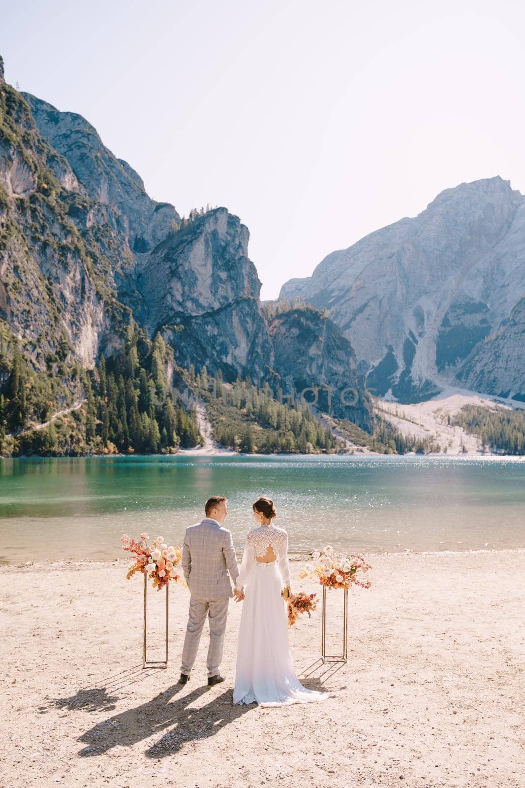 The bride and groom stand still for the ceremony, with an arch of autumn floral columns, against the backdrop of Lago di Braies in Italy. Destination wedding in Europe, at Braies lake.