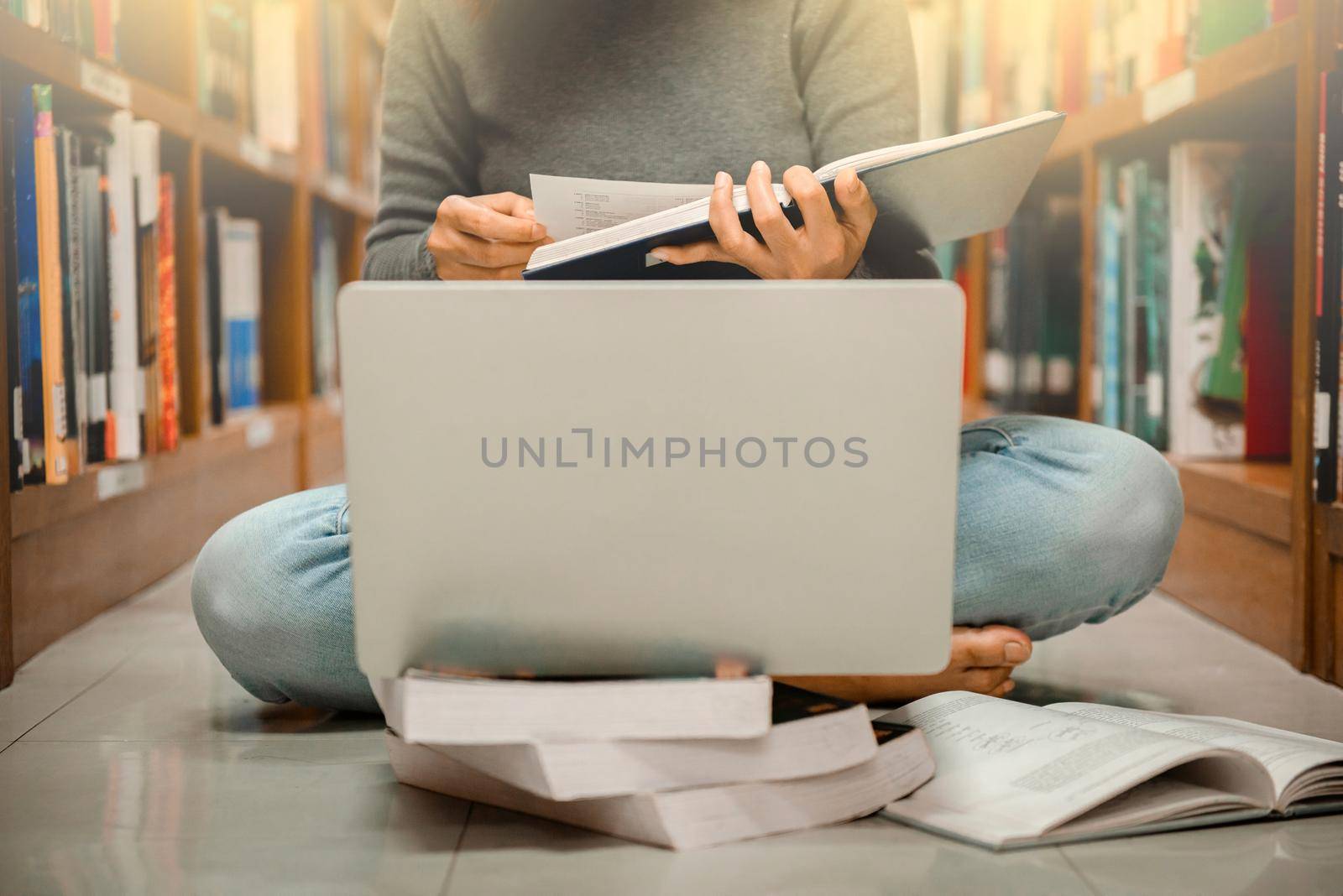 A woman sitting at library looking a book with laptop computer with blurred bookshelf background. by thanumporn