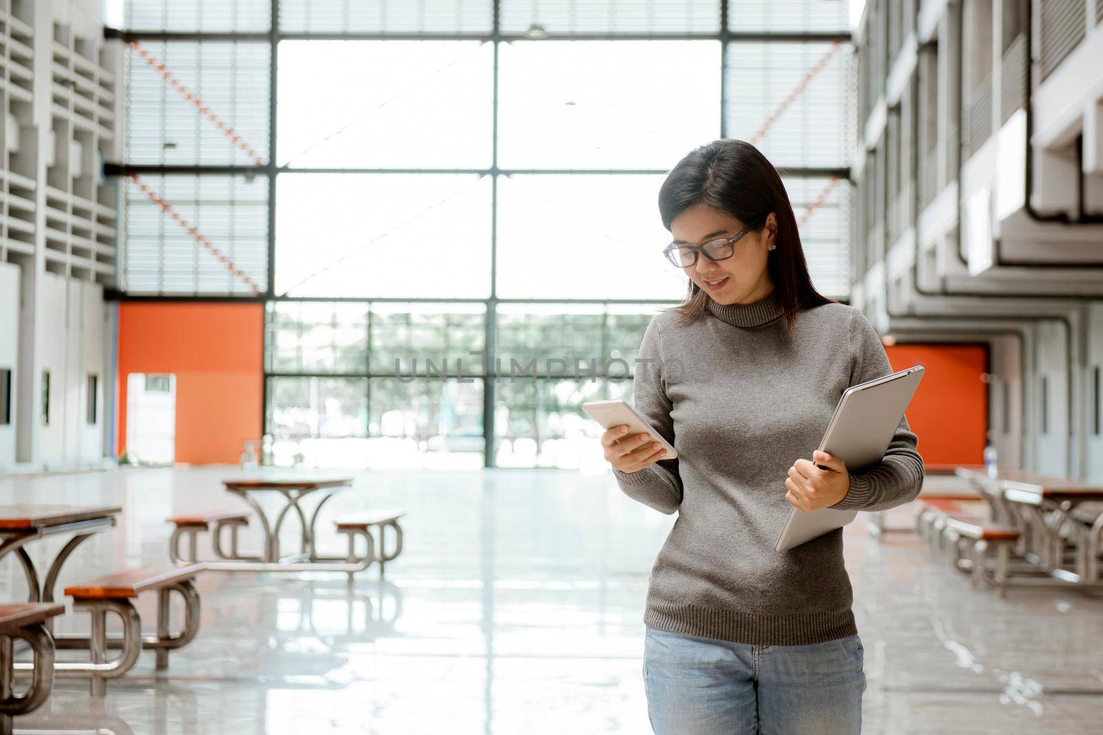 Image of young woman student walking in the building using mobile phone holding laptop computer. Chats and correspondence, online communication. by thanumporn