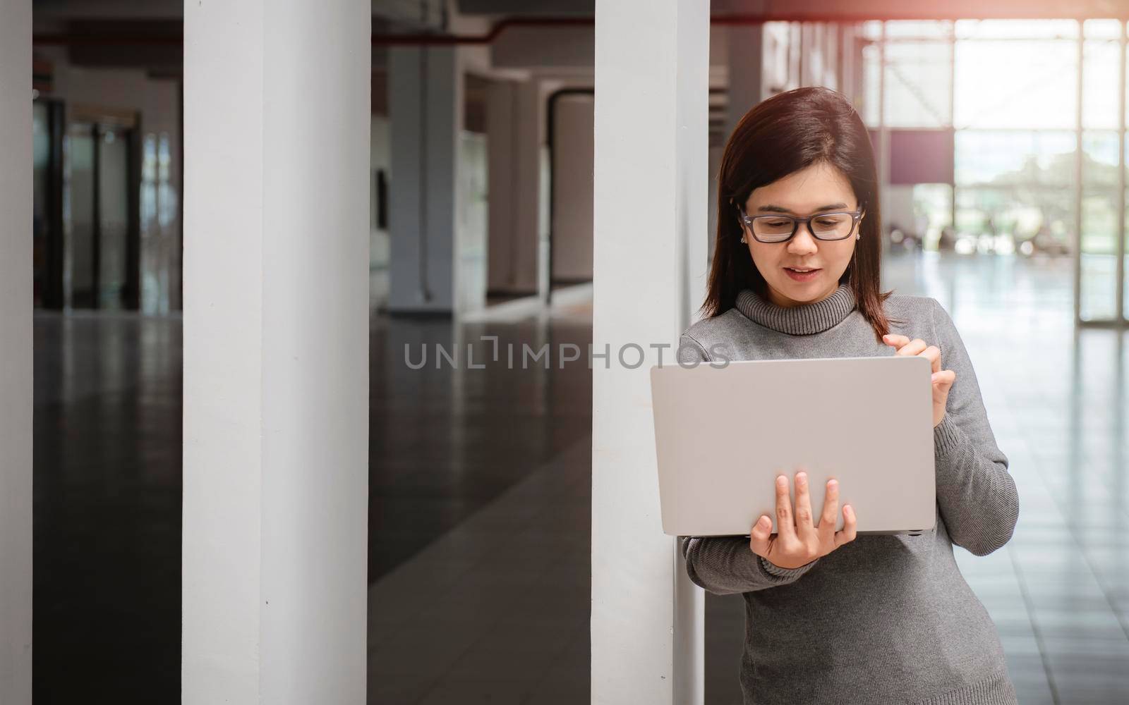 Woman in office has video conversation. Young positive woman in glasses standing in front of laptop and talking with somebody online. by thanumporn