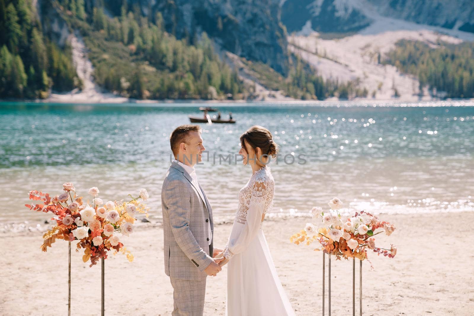 The groom dons a bride's ring, in place for the ceremony, with an arch of autumn floral columns, against the backdrop of Lago di Braies in Italy. Destination wedding in Europe, at Braies lake.
